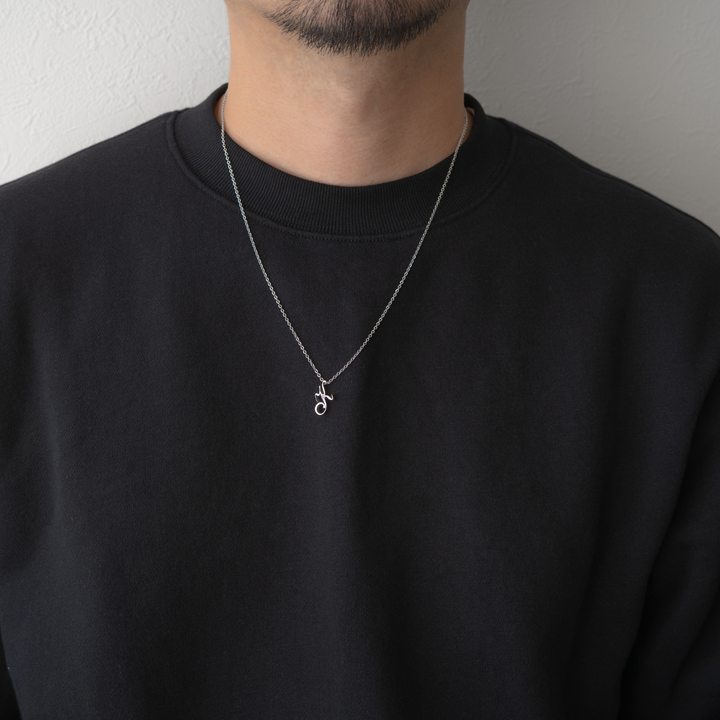 男性が黒いスウェットシャツを着て、銀色のチェーンと筆記体の「h」の文字のペンダントを身につけている