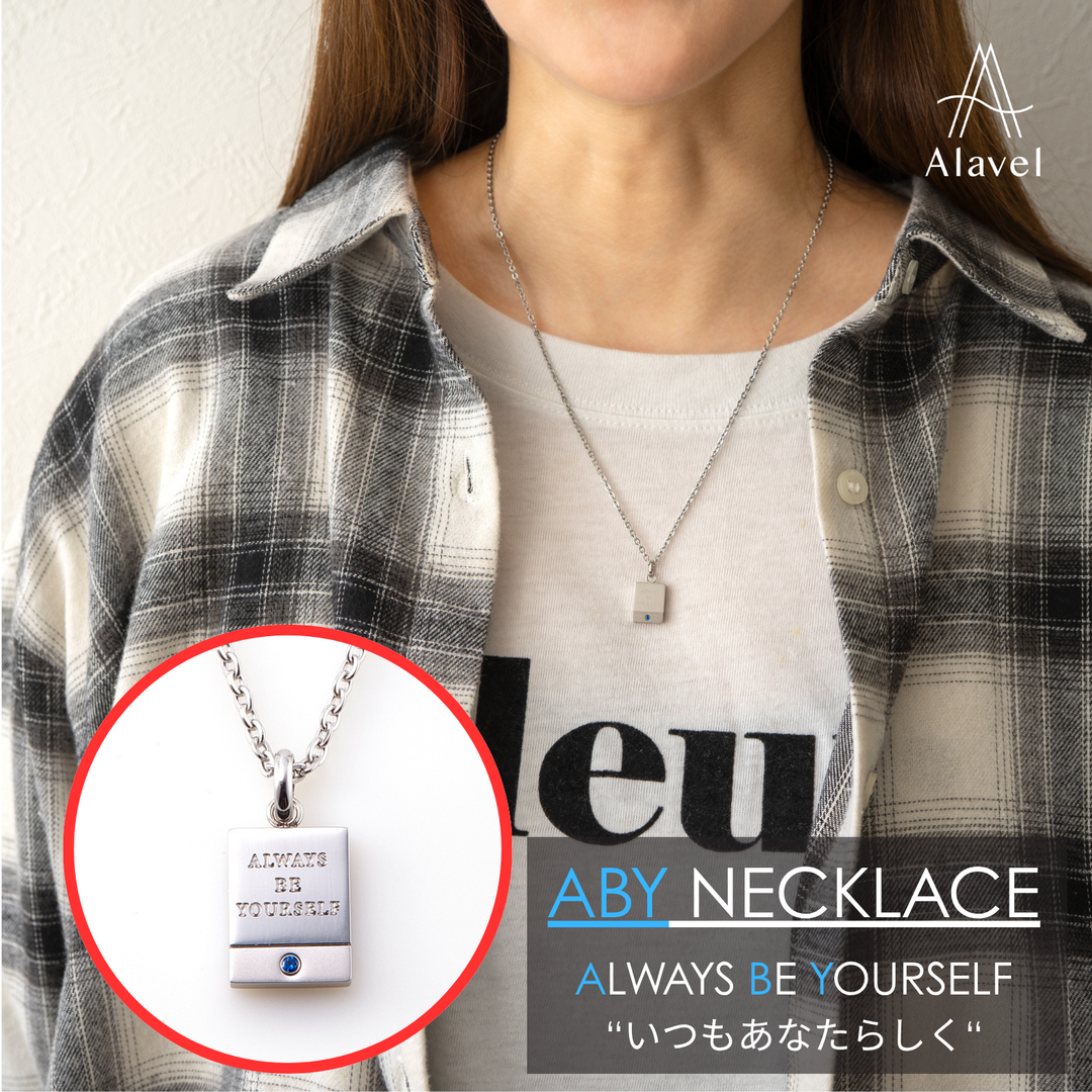 女性がチェック柄のシャツと白いTシャツを着て、Always Be Yourselfと刻印されたシルバーのネックレスを着用