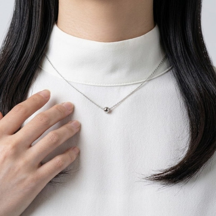 女性が白いタートルネックのシャツを着て、シンプルなシルバーのネックレスを着用し、左手を胸に当てている